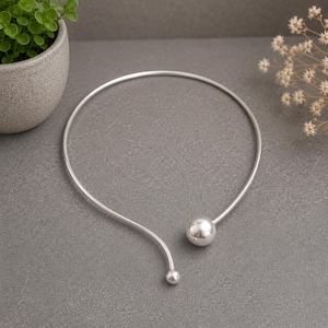 Puede incluir: Un collar rígido de metal plateado con un diseño circular abierto y liso. Un extremo del collar termina en una cuenta esférica grande y pulida, mientras que el otro extremo presenta una cuenta esférica más pequeña a juego.