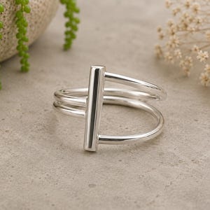 Peut inclure: Une bague en argent poli présentant un design à barre verticale reliée à un double anneau. La bague possède une finition métallique lisse et réfléchissante ainsi qu'une forme structurelle où les deux anneaux se rejoignent au centre.