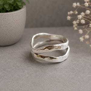 May include: A silver ring featuring a double-band design with an open, irregular, and textured surface. The metal has a polished finish with visible indentations and a wavy, organic shape that creates a layered appearance on the finger.
