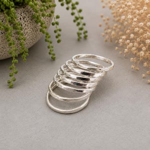 Peut inclure: Une pile de sept bagues fines en métal argenté. Chaque bague présente une texture martelée légèrement irrégulière sur la surface extérieure, créant une finition réfléchissante. Les bagues sont disposées en cascade et se chevauchent.