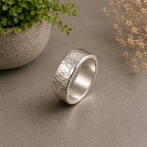 May include: A silver-colored ring with a wide band and a textured, hammered finish on the exterior surface. The interior of the band is smooth and polished, reflecting light. The ring is circular with a consistent width throughout the entire band.
