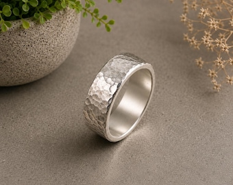 May include: A silver-colored ring with a wide band and a textured, hammered finish on the exterior surface. The interior of the band is smooth and polished, reflecting light. The ring is circular with a consistent width throughout the entire band.