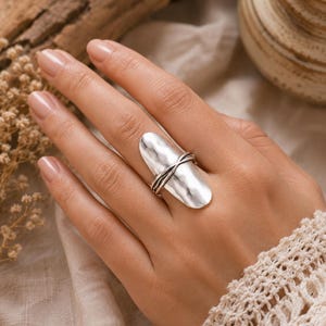 May include: A silver-toned ring featuring a large, elongated, hammered metal plate surface. The centre of the plate is accented by a thin, twisted silver wire detail that wraps horizontally around the middle, creating a layered, textured appearance.