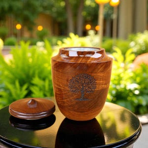 May include: A wooden urn with a tree design is displayed with its lid beside it. The urn is a warm brown color and sits on a black surface. The background is a blurred outdoor scene with greenery.