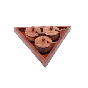 May include: A wooden spice rack with three round containers, each with a lid and a small spoon. The containers are set in a triangular wooden tray. The wood has a warm, reddish-brown tone.