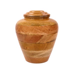 May include: A wooden urn with a lid. The urn is made of various shades of brown wood, arranged in horizontal bands. The lid is a lighter shade of brown and has a tiered design. The urn has a rounded shape.