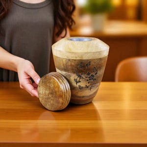 May include: A wooden urn with a lid, featuring a natural wood grain finish and a decorative hummingbird and butterfly design. The urn is cylindrical with a light-colored top and a darker, banded lower section.