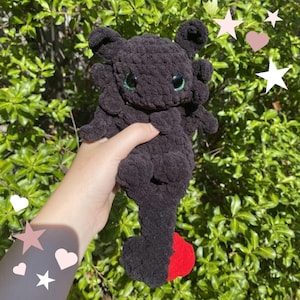 May include: A dark charcoal grey crocheted plush toy featuring a rounded head with large black eyes, small ears, and a long tail ending in a bright red tip. The soft, textured yarn construction gives the creature a fuzzy appearance and tactile feel.