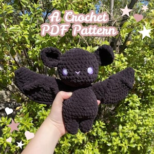 Może przedstawiać: Czarny szydełkowy nietoperz z dużymi skrzydłami, uszami i fioletowymi oczami. Tekst "A Crochet PDF Pattern" jest wyświetlany w kolorze różowym. Nietoperz jest trzymany na tle zielonego, liściastego tła.