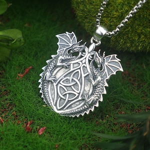 Peut inclure: Un collier pendentif argenté avec un motif de dragon détaillé. Le pendentif présente deux dragons encadrant un nœud celtique, avec des détails d'ailes et d'écailles. Le collier est présenté sur de la mousse et de l'herbe vertes.