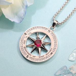 May include: A silver-toned compass pendant necklace featuring a central round red gemstone, a star-shaped compass rose, and a circular frame set with small clear stones. The outer ring is engraved with the text "in the direction of your dreams".