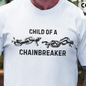 May include: White t-shirt with the text "CHILD OF A CHAINBREAKER" in black, featuring a chain design. The shirt is short-sleeved and has a crew neck. The image also shows a dark blue tumbler.