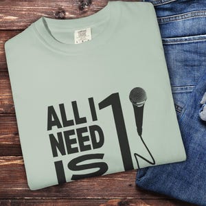 May include: A light green t-shirt with the text "ALL I NEED IS 1" in black, along with a microphone graphic. The shirt is folded on a wooden surface next to a pair of blue jeans.