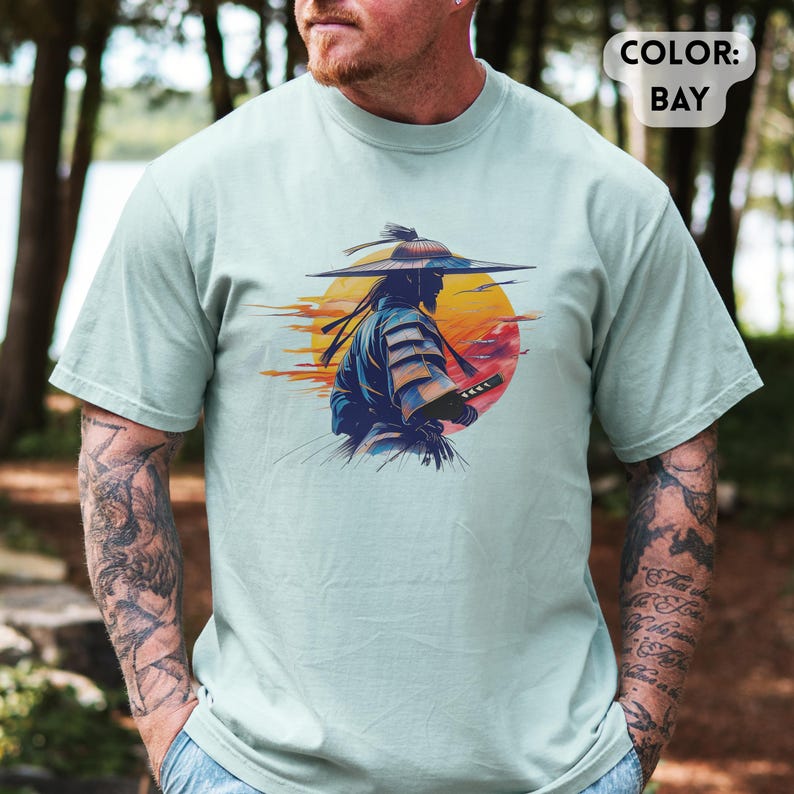 Op de afbeelding: Lichtgroen t-shirt met een afbeelding van een samoerai-krijger met een zwaard, tegen een zonsondergang. De samoerai is afgebeeld in blauw en goud, met een grote hoed. De tekst "COLOR: BAY" staat rechtsboven.