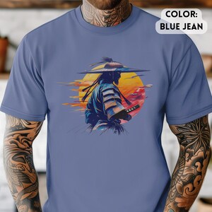 Op de afbeelding: Blauw spijkerbroekkleurig T-shirt met een afbeelding van een samoerai-krijger met een zwaard, tegen een zonsondergang. De samoerai is afgebeeld als silhouet, gekleed in traditionele kleding en een brede hoed. De zonsondergang bevat tinten geel, oranje en rood.