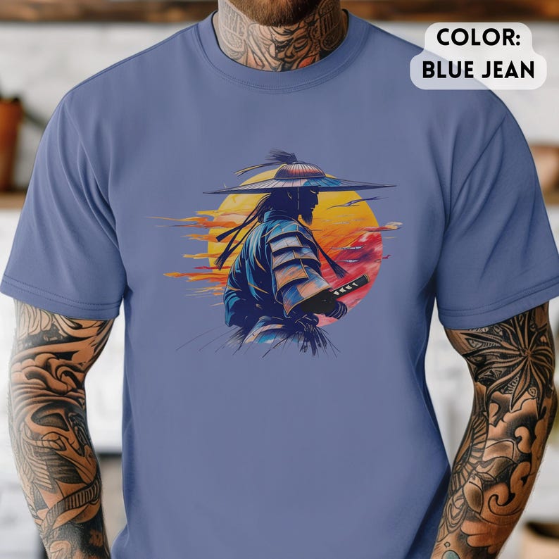 Op de afbeelding: Blauw spijkerbroekkleurig T-shirt met een afbeelding van een samoerai-krijger met een zwaard, tegen een zonsondergang. De samoerai is afgebeeld als silhouet, gekleed in traditionele kleding en een brede hoed. De zonsondergang bevat tinten geel, oranje en rood.