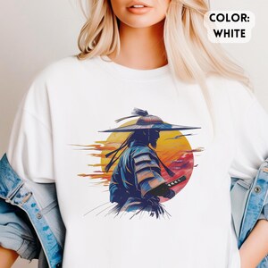 Op de afbeelding: Wit T-shirt met een afbeelding van een samoerai-krijger met een zwaard, tegen een zonsondergang. Het ontwerp gebruikt blauw, oranje en geel. De tekst "COLOR: WHITE" wordt weergegeven in een wit kader.