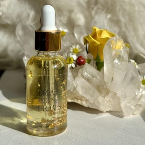 May include: A clear glass bottle with a gold cap and dropper, filled with a golden liquid and gold flakes. The bottle has the text "DREAM HOUSE HERBALS" printed on it. The background includes a yellow rose, crystals, and small white flowers.