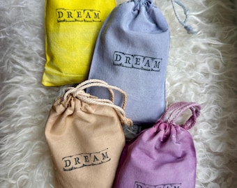 May include: Four small fabric drawstring pouches in yellow, light blue, beige, and purple. Each pouch features the word DREAM printed in a black, rectangular stamp-style font on the front and includes a matching drawstring closure.
