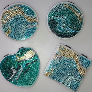 May include: Four compact mirrors in different shapes: round, heart, and square. Each mirror is decorated with a pattern of teal, gold, and clear rhinestones. The mirrors have a white border and a small black hinge.