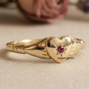 May include: A gold-colored ring with a heart-shaped centerpiece. The heart features a small, red gemstone at its center, surrounded by a star design. The ring has a delicate, detailed design, with the text "sagat gems" visible on the heart.