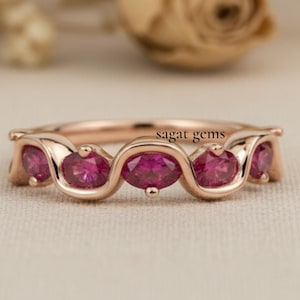 May include: Rose gold ring with five oval-cut, vibrant pink gemstones. The ring has a delicate, wavy design. The text "sagat gems" is visible on the ring. The background features blurred flowers.