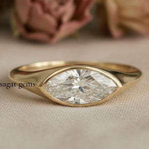 Könnte beinhalten: Ein goldener Ring mit einem Diamanten im Marquiseschliff. Der Diamant ist in einer Zargenfassung gefasst. Der Ring liegt auf einer neutralen Oberfläche mit verschwommenen Blumen im Hintergrund. Der Text "sagat gems" ist sichtbar.