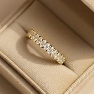 Könnte beinhalten: Ein goldener Ring mit einer Reihe oval geschliffener Edelsteine liegt in einer offenen, beigefarbenen Samtringbox. Die Ringbox trägt den Schriftzug "Sagat Gems". Die Edelsteine des Rings werden prominent präsentiert.