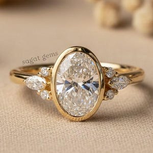 Könnte beinhalten: Ein goldener Ring mit einem oval geschliffenen Diamanten als Mittelstein. Der Ring hat kleinere Akzentsteine auf beiden Seiten des Mittelsteins. Der Ring ist auf einem neutralen Hintergrund platziert. Der Text "sagat gems" ist sichtbar.