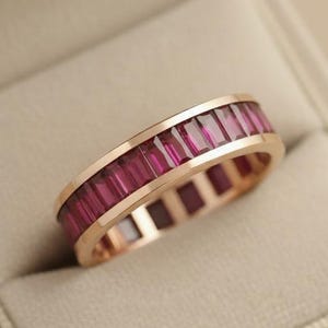 Può includere: Un anello in oro rosa con una fascia di pietre preziose rettangolari rosa intenso. L'anello è presentato in un portagioie di velluto beige chiaro, su uno sfondo morbido e neutro.