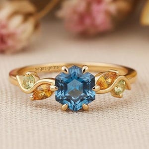 May include: A gold ring featuring a hexagonal, blue gemstone as the centerpiece. The ring is adorned with smaller, leaf-shaped gemstones in shades of yellow and green. The ring is set against a soft, neutral background with blurred floral elements.