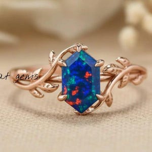 Op de afbeelding: Roségouden ring met een zeshoekige, donkerblauwe opaal met rode en groene vlekken. De ring heeft een delicaat bladerontwerp op de band. De tekst "sagat gems" is zichtbaar op de band.