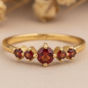 Puede incluir: Un anillo de oro con cinco piedras rojas. La piedra central es más grande que las otras. El anillo está sobre una superficie de color neutro, con el texto "sagat gems" visible.