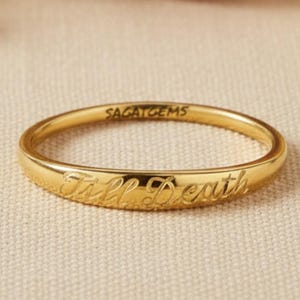 Könnte beinhalten: Ein goldfarbener Ring mit der Inschrift "Till Death" auf dem Band. Der Ring ist dünn und liegt auf einer neutralfarbenen Oberfläche. Der Markenname "SAGATGEMS" ist ebenfalls auf dem Ring eingraviert.