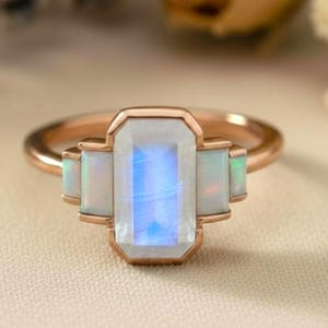 May include: A rose gold ring with a rectangular moonstone centre stone, displaying an iridescent effect, and two smaller rectangular opal stones on either side. The ring is set against a neutral background with floral accents.