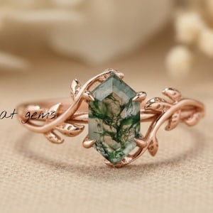 Op de afbeelding: Een roségouden ring met een zeshoekige mosagaat edelsteen. De ringband heeft blad- en wijnstokdetails. De edelsteen is lichtgroen met donkerdere groene insluitsels. Sieraden.