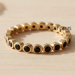 May include: A gold ring featuring a continuous line of small, round, black gemstones. The ring is displayed on a light-coloured surface. The text "SAGAT GEMS" is visible on the ring.