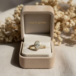 May include: A gold ring with an oval-shaped diamond in a velvet ring box. The ring has a detailed band and a halo of smaller diamonds around the center stone. The ring box is beige, and the text "sagat gems" is printed on the inside of the box.