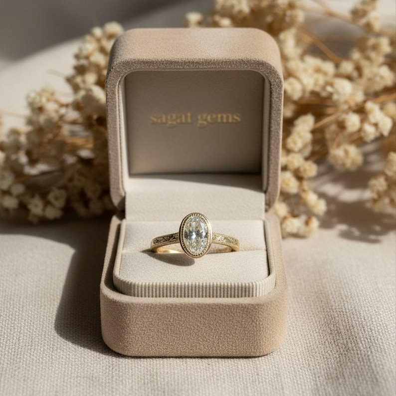 May include: A gold ring with an oval-shaped diamond in a velvet ring box. The ring has a detailed band and a halo of smaller diamonds around the center stone. The ring box is beige, and the text "sagat gems" is printed on the inside of the box.