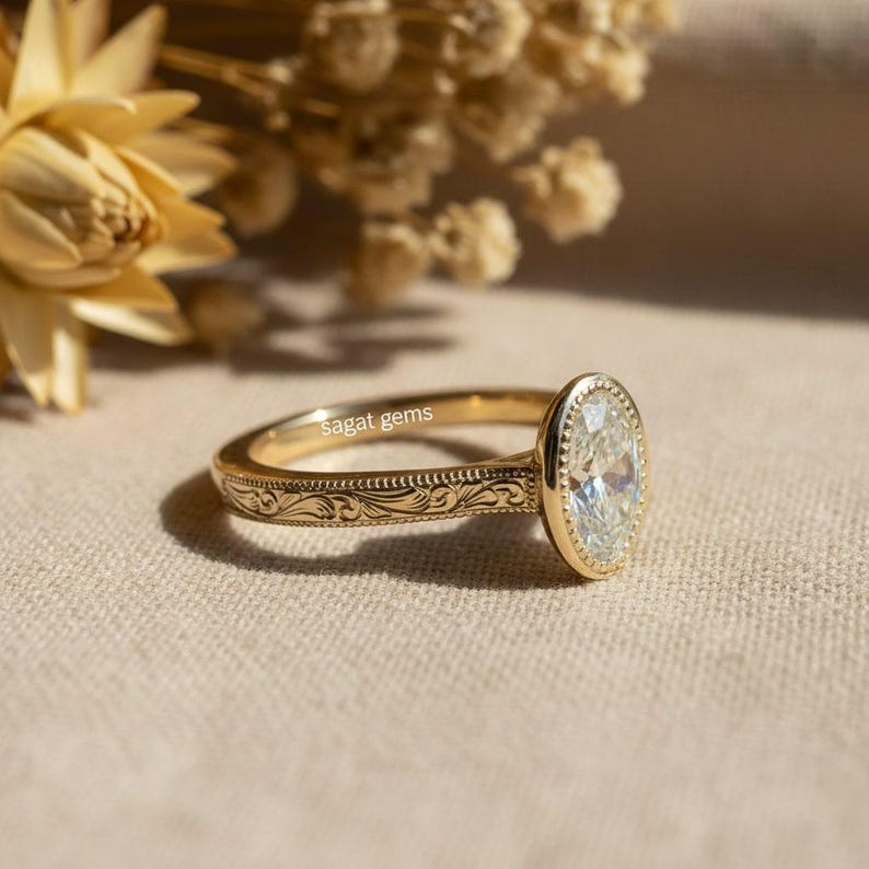 May include: A gold ring with an oval-shaped, clear gemstone. The ring band features an engraved floral pattern. The ring is set against a neutral background with dried flowers in the upper left corner. The text "sagat gems" is visible on the ring.