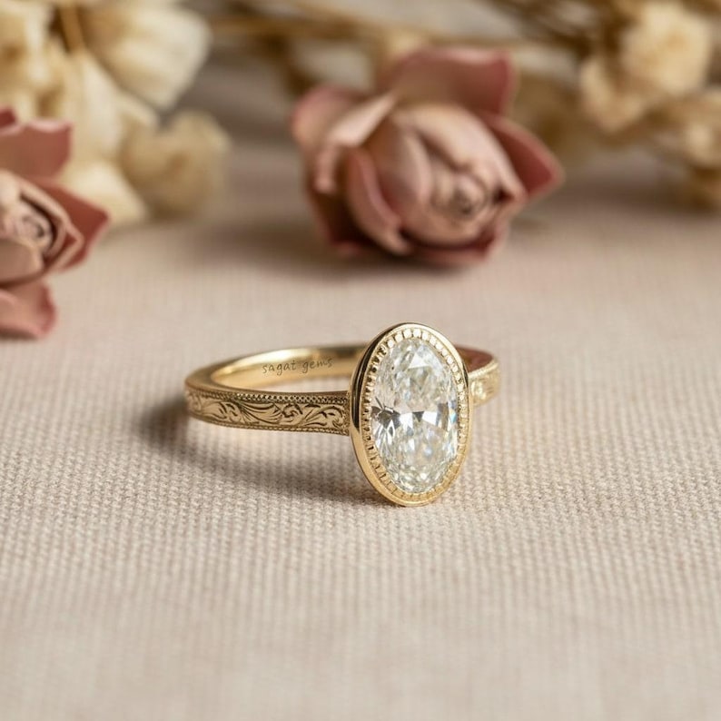 May include: A gold engagement ring with an oval-cut diamond. The ring has a detailed band with an engraved pattern. The diamond is set in a bezel setting. The ring is displayed on a neutral-colored surface with blurred flowers in the background.