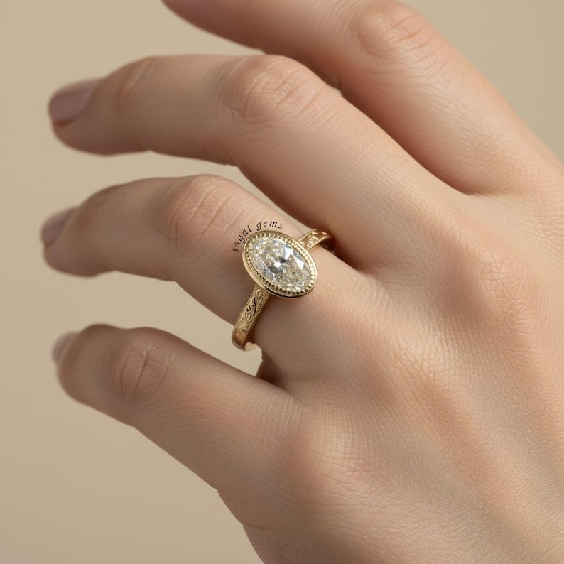 May include: A gold ring with an oval-shaped, clear gemstone. The ring has a detailed, engraved band and a bezel setting. The ring is on a finger, with the hand in the frame. The ring is from "sagal gems".