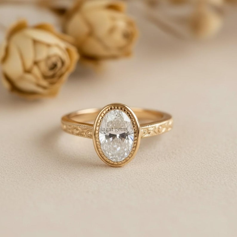 May include: A gold ring with an oval-shaped, clear gemstone. The ring has a detailed, engraved band and a bezel setting around the gemstone. The text "sagat gems" is visible on the gemstone. The background features dried flowers.