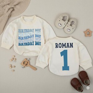 May include: Cream-colored baby bodysuit with the text "BIRTHDAY BOY" in blue. The back of the bodysuit has the name "ROMAN" and the number "1" in blue. Brown leather baby shoes and bear-shaped slippers are also in the image.