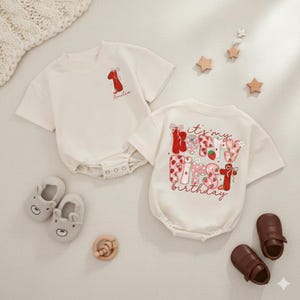 May include: Two cream-colored baby onesies. One has a red number one graphic with the name "Amelia". The other has the text "it's my FIRST birthday" in a strawberry and floral design. Includes baby shoes and a wooden toy.