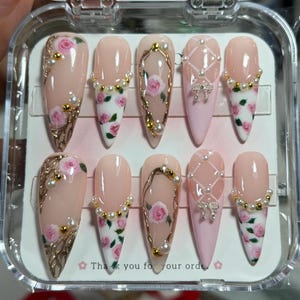 May include: A set of ten almond-shaped press-on nails in a clear plastic case. The designs feature a nude and light pink base with hand-painted pink roses, green leaves, gold metallic accents, small white pearls, and silver-toned bow charms.