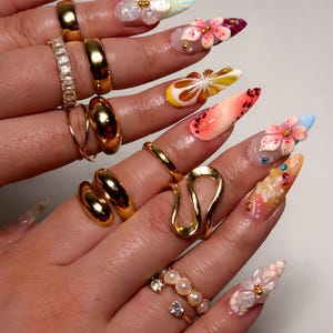 May include: A hand displays a collection of gold rings and long, decorated fingernails. The rings include bands, a double ring, and a unique open design. The nails feature floral and abstract designs with rhinestones and pearls.