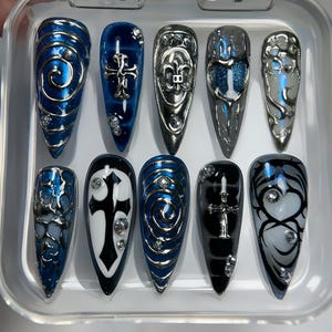 May include: A set of ten long, pointed false nails in a clear plastic case. The nails feature various designs, including swirls, crosses, and ornate silver accents. Colors include blue, silver, black, and white, with some nails adorned with rhinestones.