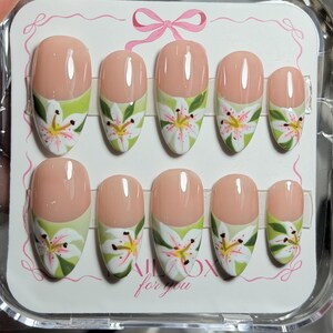 May include: A set of ten press-on nails in a clear plastic case, featuring a nude base with a light green French tip and a hand-painted white lily floral design with yellow and pink accents. The display card includes a pink bow and the text NAILBOX for you.