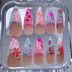 May include: A set of ten artificial nails in a clear plastic case. Each nail is almond-shaped and features a floral design in shades of pink, blue, and white. The base of each nail has a natural, nude color.